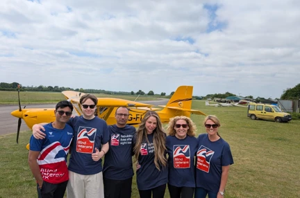 All six of Team Thea stood in front of a small yellow plane which is sat on a grassy field with a runway behind. The team are all wearing Blind Veterans UK T-shirts.