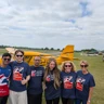 All six of Team Thea stood in front of a small yellow plane which is sat on a grassy field with a runway behind. The team are all wearing Blind Veterans UK T-shirts.