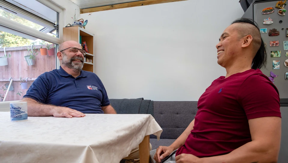 Blind veteran Ken and his support worker sitting at a table laughing together in Ken's home