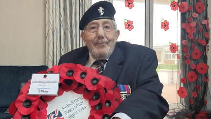 Pat is sat in his wheelchair wearing his medals and holding the wreath. Behind him is a curtain of poppies
