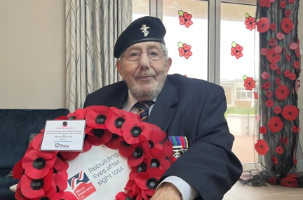 Pat is sat in his wheelchair wearing his medals and holding the wreath. Behind him is a curtain of poppies