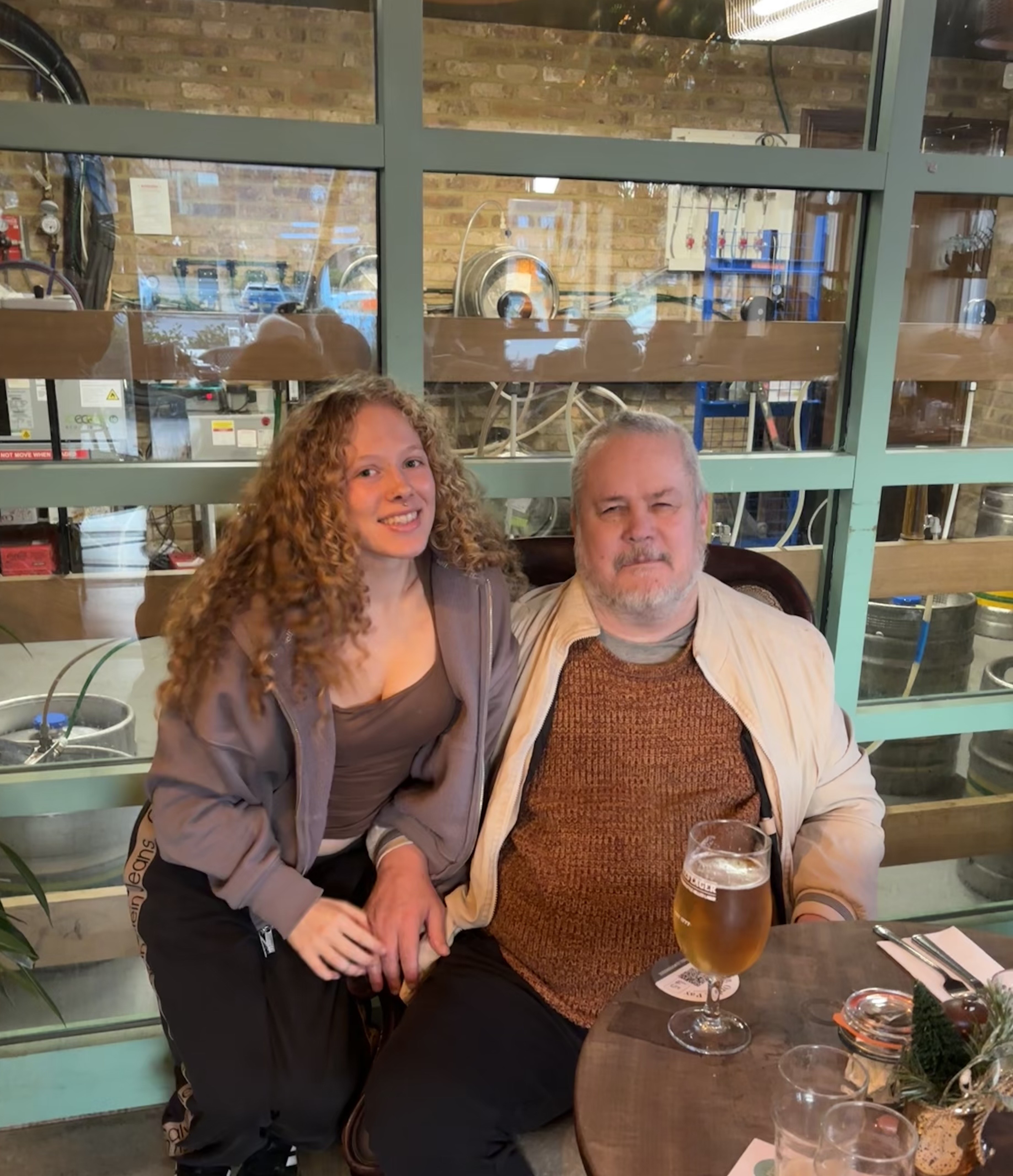 Anna and Mark sat side by side at a table in the pub. They are holding hands and smiling at the camera.