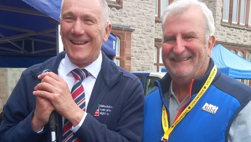 Blind veteran John is holding a cane and smiling as he stands next to volunteer Derek