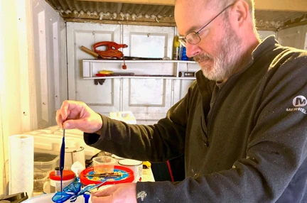 Graham holding a resin butterfly over a bucket while pouring blue paint over the top