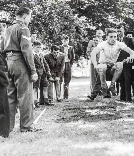 A blind veteran jumping mid-air during an outdoor sports event, surrounded by other veterans, staff and onlookers.