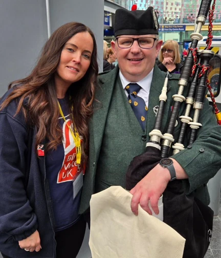 A Blind Veterans UK staff member stands next to a man in uniform holding bagpipes.