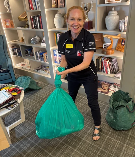 A lady with long red hair, wearing a navy blue t-shirt and trousers branded with the Blind Veterans UK logo stands looking at the camera and smiling. She is holding a yellow bin bag that is full of rubbish and tied in a knot at the top.