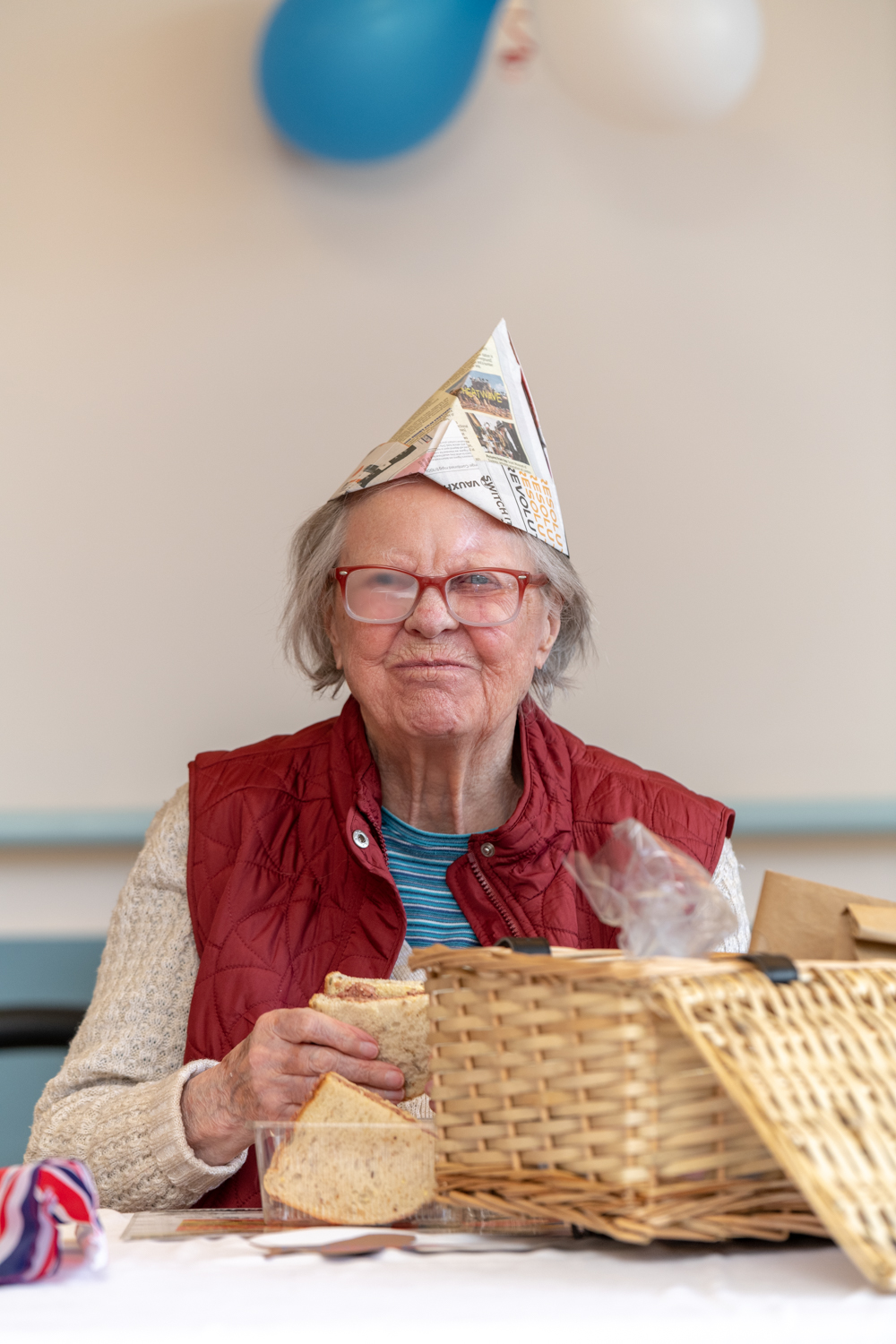 Nancy is sat at a table wearing a hat made from folded newspaper and eating sandwiches from a hamper