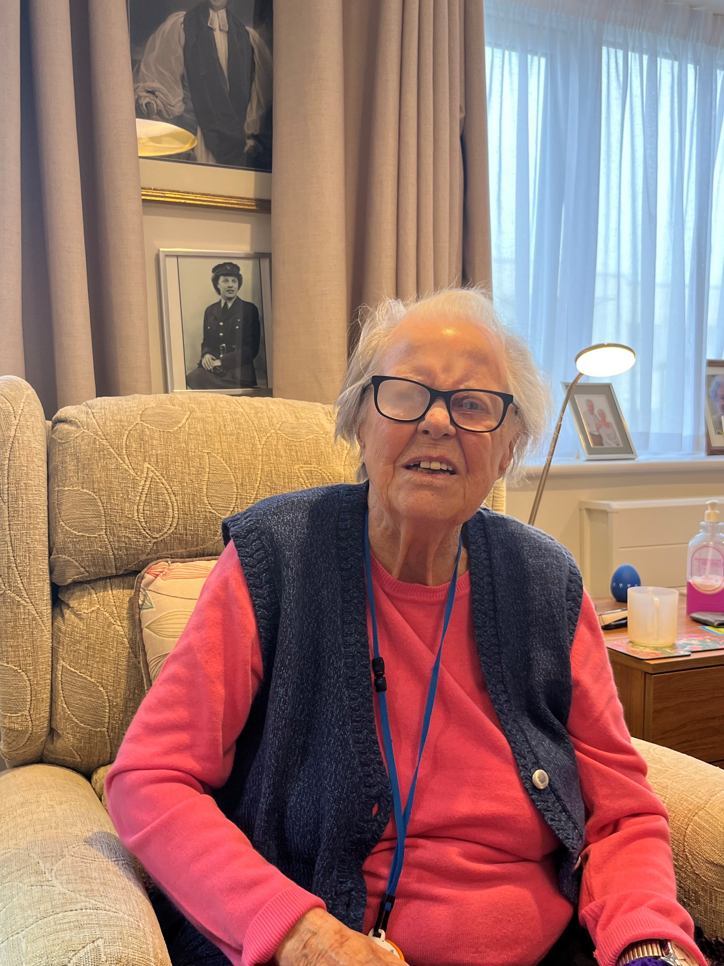 Nancy is sat in an armchair with a picture of her during her service hanging on the wall behind