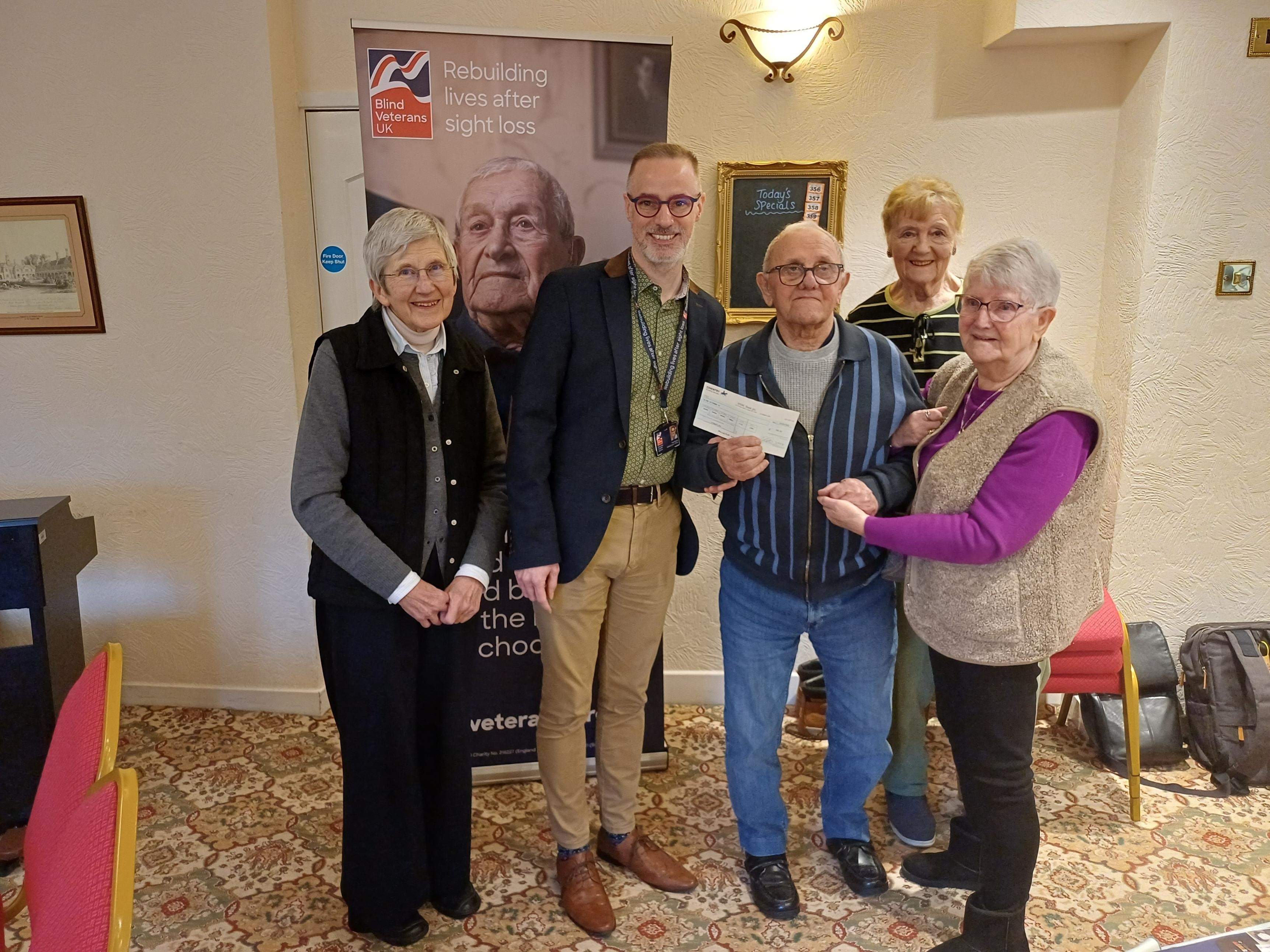 A group of five people with Ray and Paul stood in the centre holding a cheque. The group are stood in front of a Blind Veterans UK pull up banner