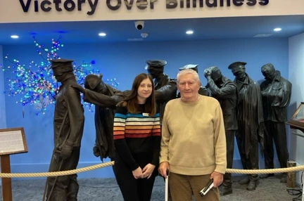 Stephanie and Mike are stood together in front of the 'Victory over Blindness' sculpture depicting a group of soldiers from the First World War leading one another away from the battlefield. Mike is holding hi white cane.