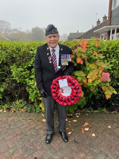 Ronald stands in his front garden smartly dressed and displaying his medals. He holds his wreath.