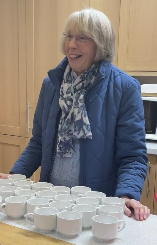 A lady stand behind a counter laid out with tea cups