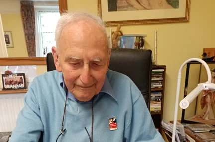 Blind veteran John, sitting at a desk with a large notepad, pen in hand, writing his memoirs