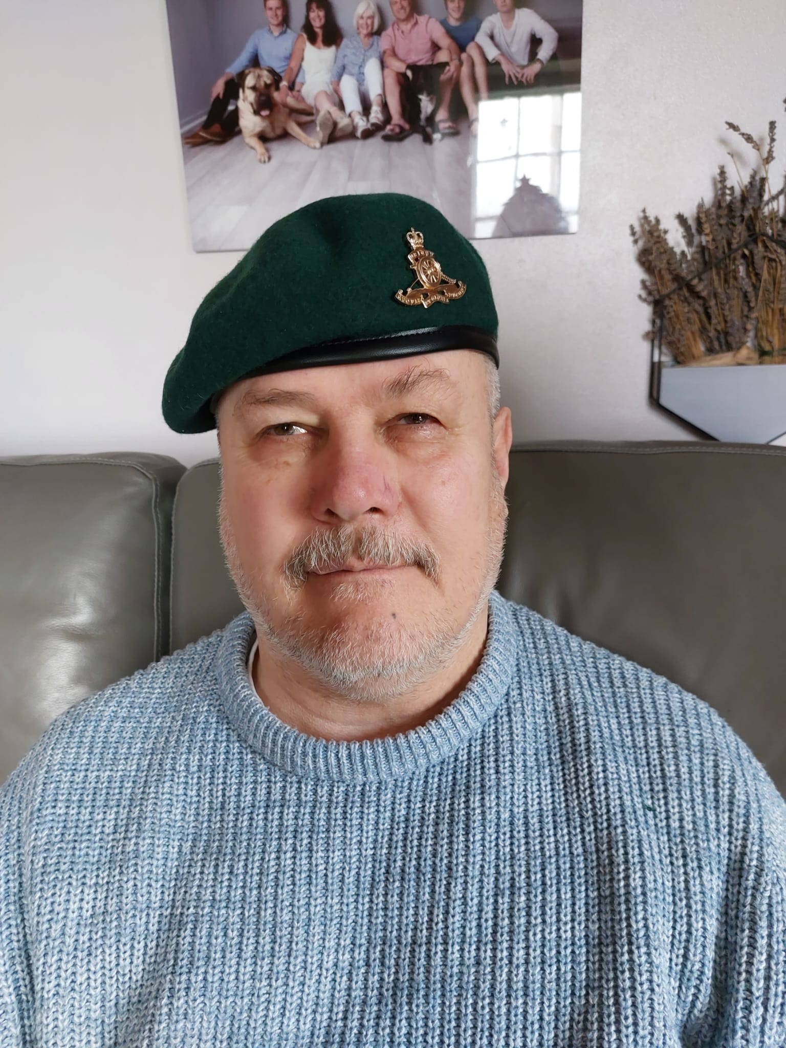 Mark sits on the sofa with a family photo behind him. He wears an Army beret and a blue knitted jumper.