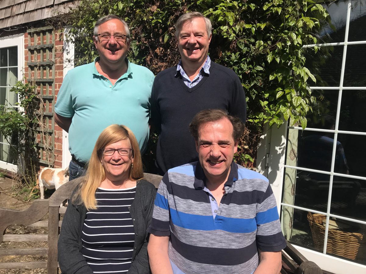 Linda and Nigel are sat on a wooden bench in the sun and Andy and Jerry are stood behind them