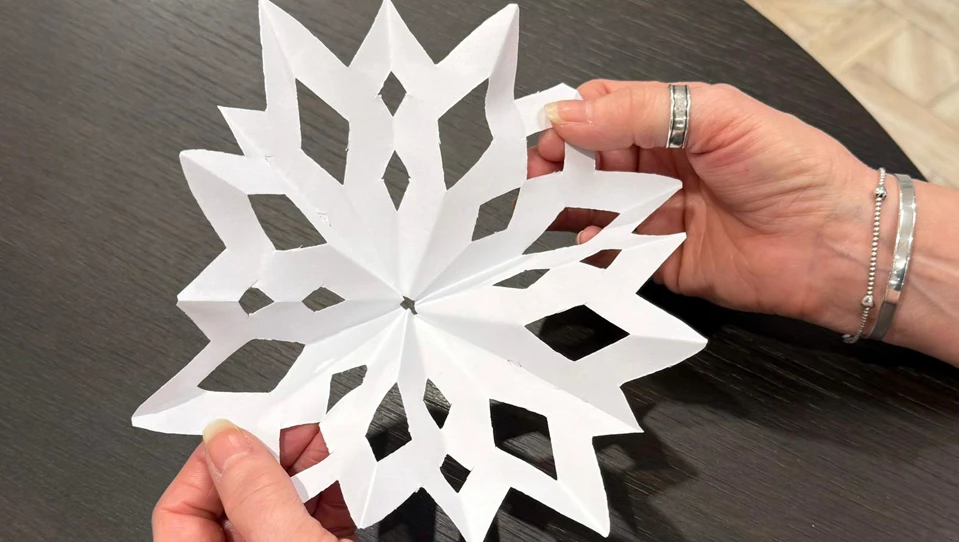 he image shows a person holding up an unfolded paper snowflake, revealing its intricate design. The snowflake has a symmetrical, decorative pattern, and the hands gently display the finished craft against a dark table background.