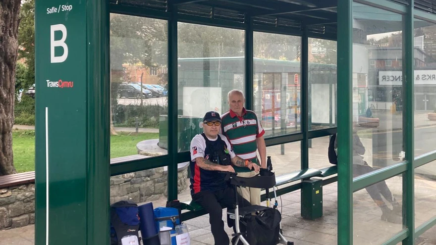 Tony and Dai sat in a bus shelter with their rucksacks beside them. Tony has his walker and is wearing his Blind Veterans UK t-shirt