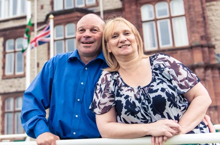 Blind veteran Billy with his arm around his wife, smiling as they stand outside our Centre of wellbeing in Llandudno