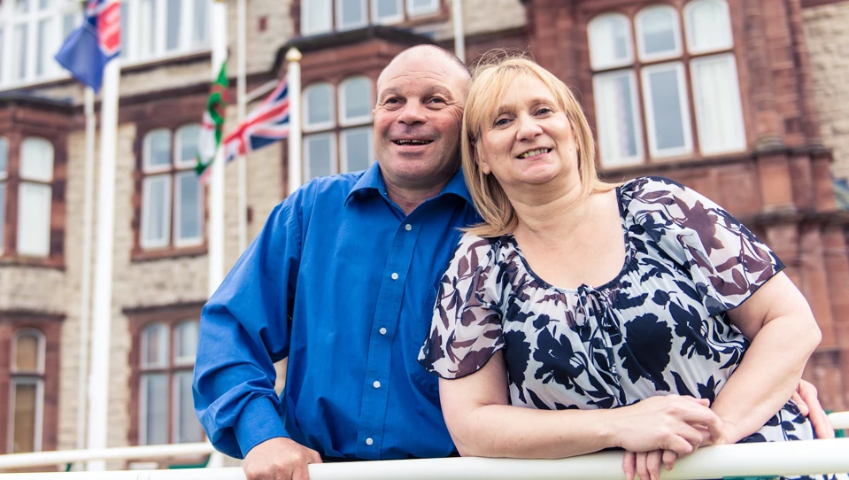 Blind veteran Billy with his arm around his wife, smiling as they stand outside our Centre of wellbeing in Llandudno