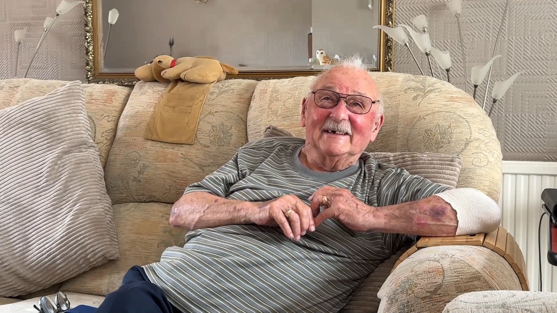 Ken sits on a sofa in his living room, smiling, with his hands resting together in front of him.