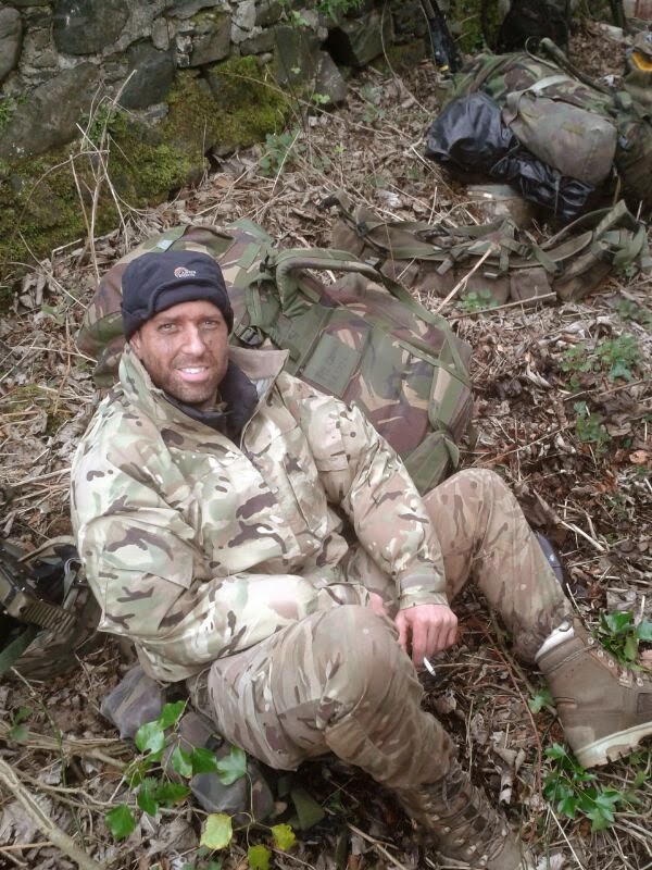 A young Simon sitting on the ground in a wooded area wearing camouflage clothing 