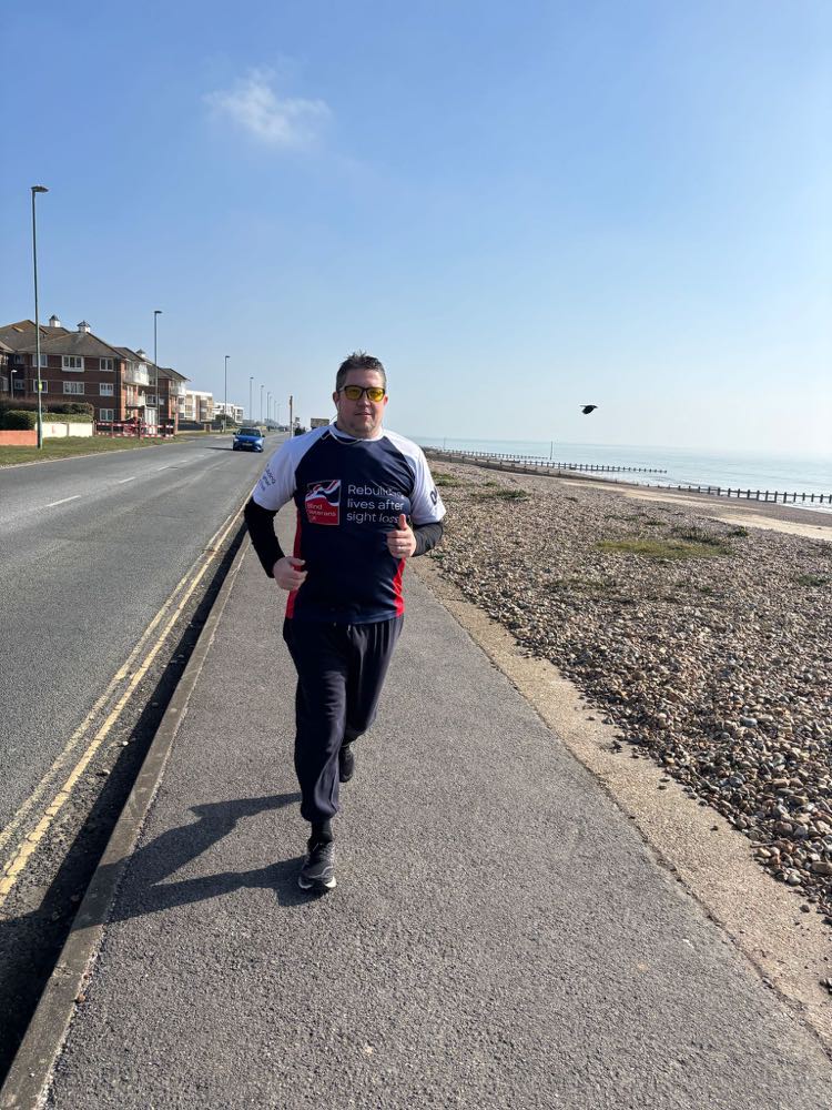 Wayne wearing his Blind Veterans UK t-shirt and tinted yellow glasses running alongside the pebble beach