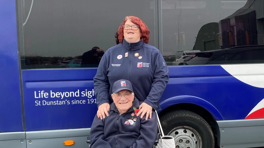 Paula stood behind blind veteran Bill. Bill is sat in his wheelchair and Paula has he hands on his shoulders.  Both are smiling for the camera.