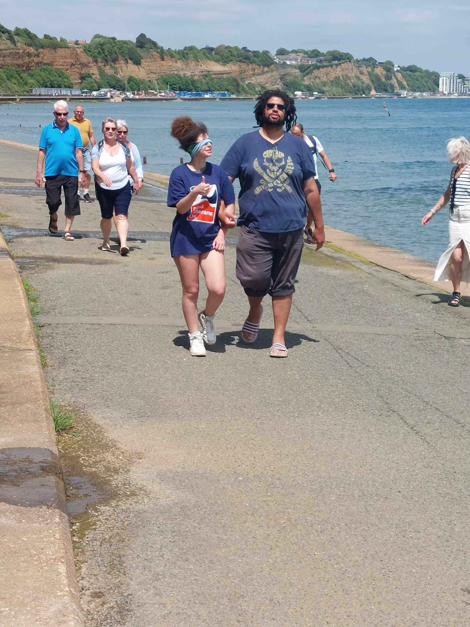 A lady in a Blind Veterans UK t-shirt is blindfolded as she walks along a promenade with the sea alongside her. She has her arm linked with a man who is guiding her