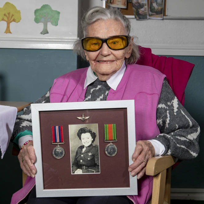 Neena smiling while sat in a chair holding a framed photo of herself in service alongside her medals