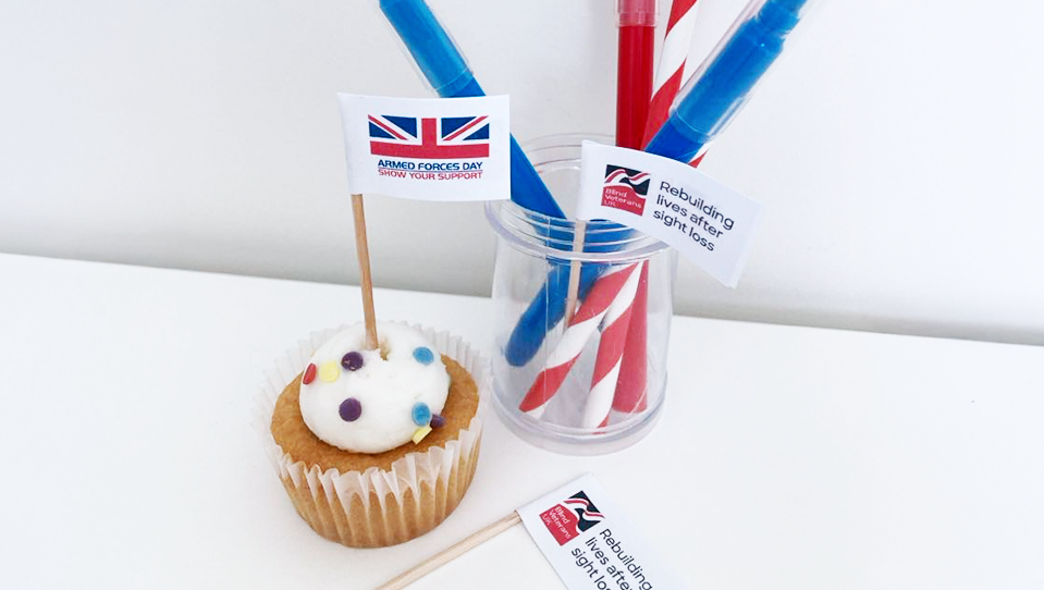 An iced mini cup cake with colourful sprinkles and a cake topper showing the National Armed Forces Day Logo, along side craft materials.