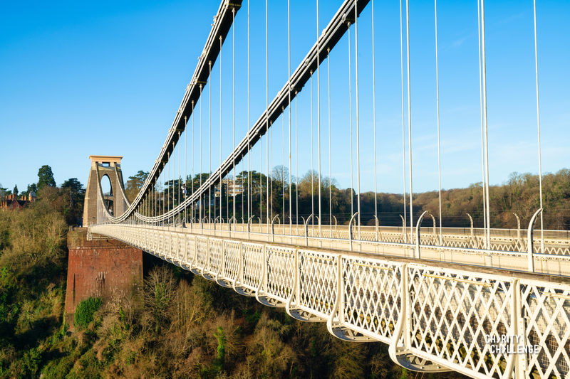 Clifton Suspension Bridge stretching away from the camera and over tall trees below it, with a bright blue sky above.