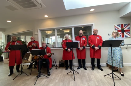 Six Chelsea Pensioners are lined up and singing. They are all wearing their famous red jackets. One is playing the guitar and two are playing the ukulele