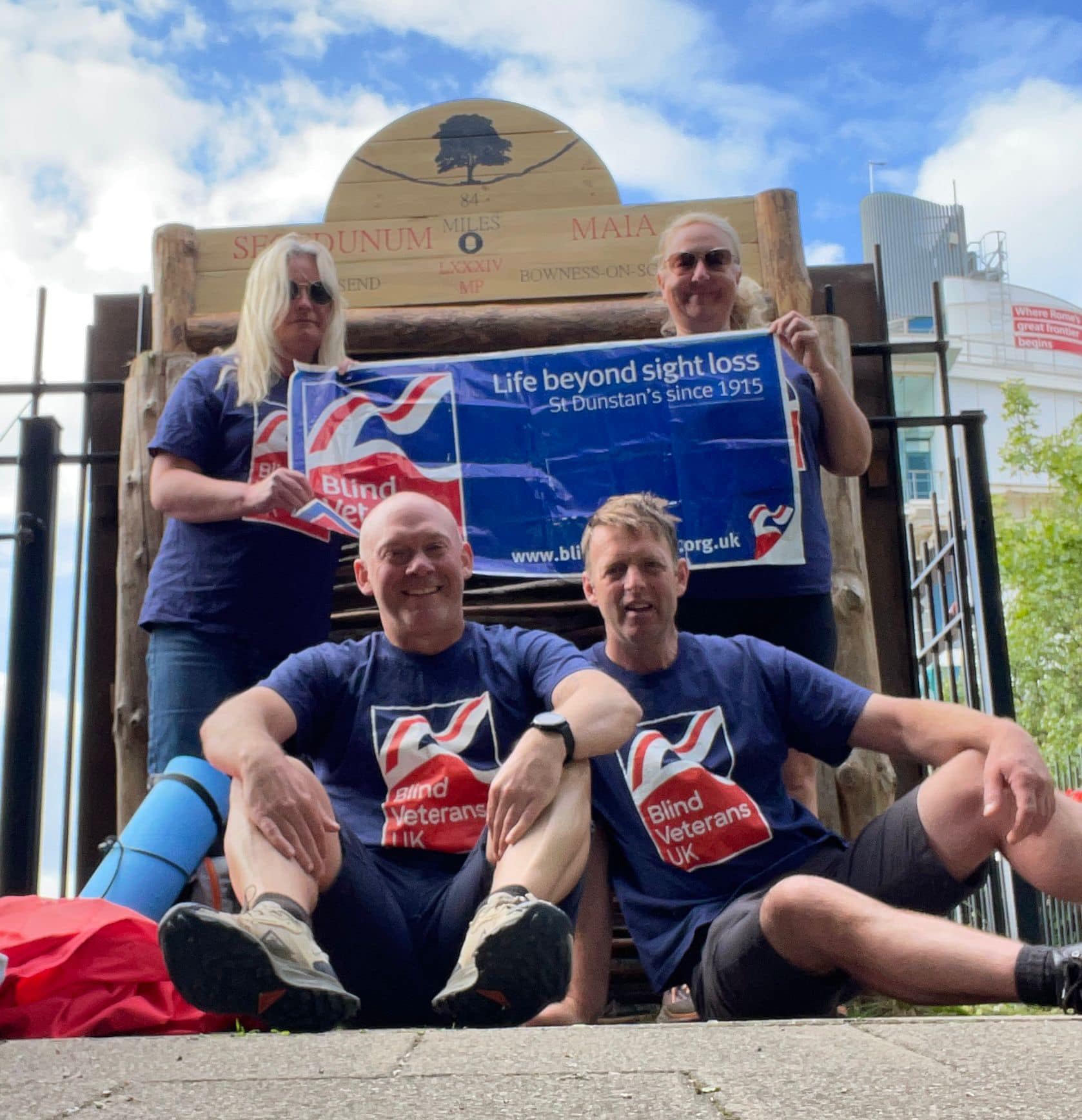 Janine and three others posing with the sign that signals they have completed the length of Hadrian's Wall. They are all in their Blind Veterans UK t-shirts and holding up a Blind Veterans UK flag