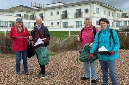 A group of four people wearing waterproof coats stand in a line on a pebble beach. They are looking at the camera and holding litter pickers and bags.