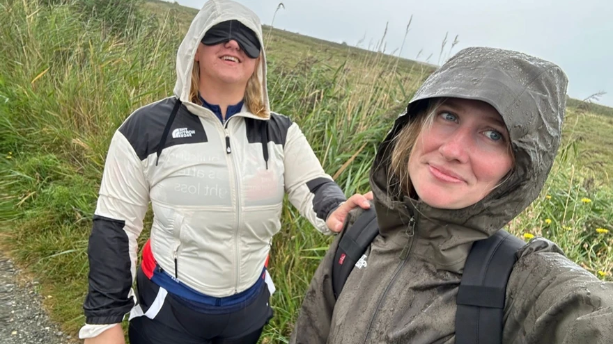 Kat is blindfolded and had her hand on Rosie's shoulder as they stop to pose for a photo. Both women are wearing rain coats with their hoods up and it is raining