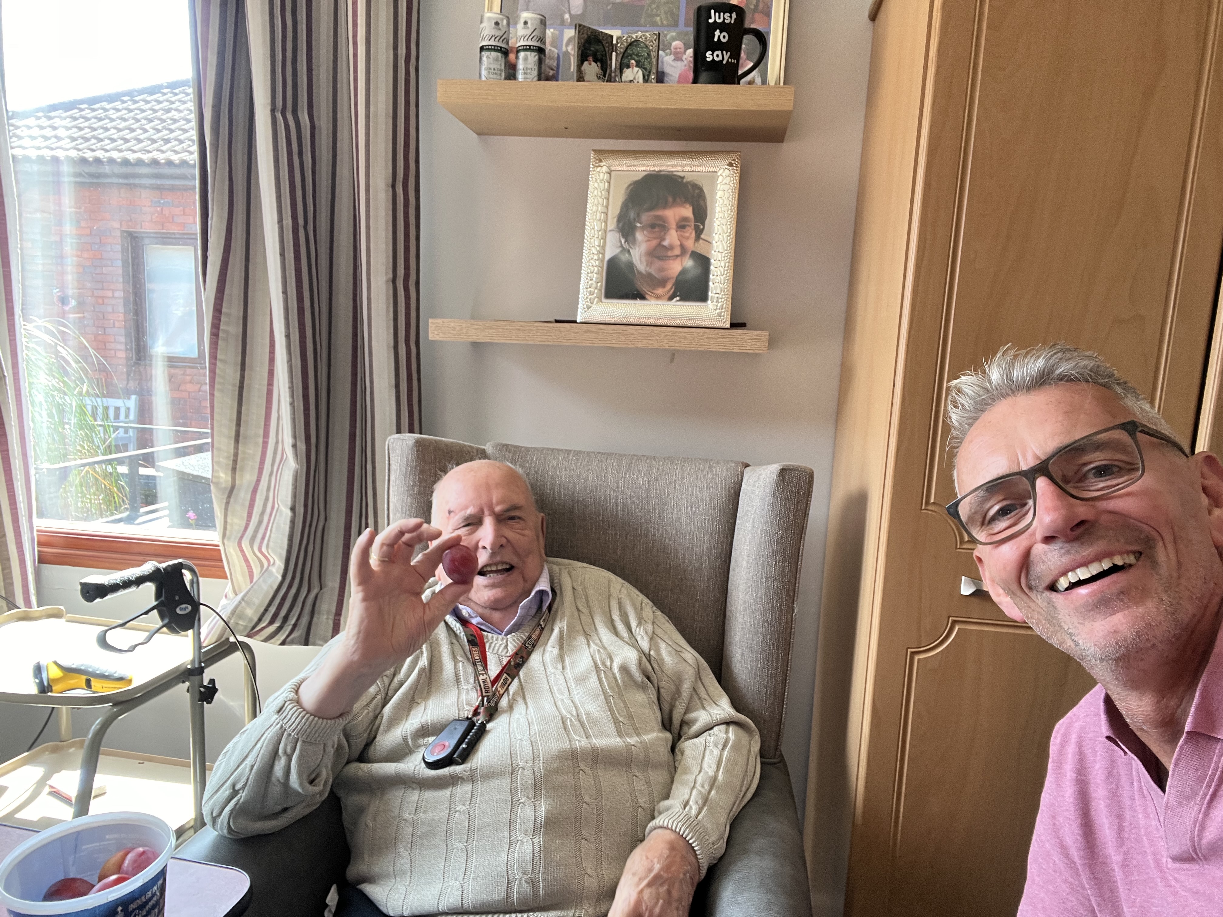 An older man sits in an armchair holding up a plum, a younger man on the right of the screen is looking at the camera smiling as he takes a selfie of them both.