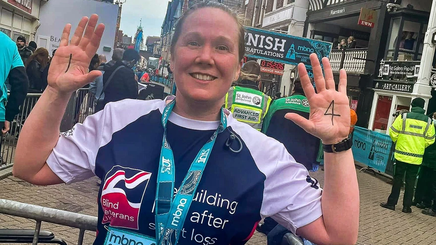 Supporter Alison wearing her marathon medal and Blind Veterans UK training top, holding up her hands to show her palms, with the number 1 written on her left palm, and 4 on her right palm.