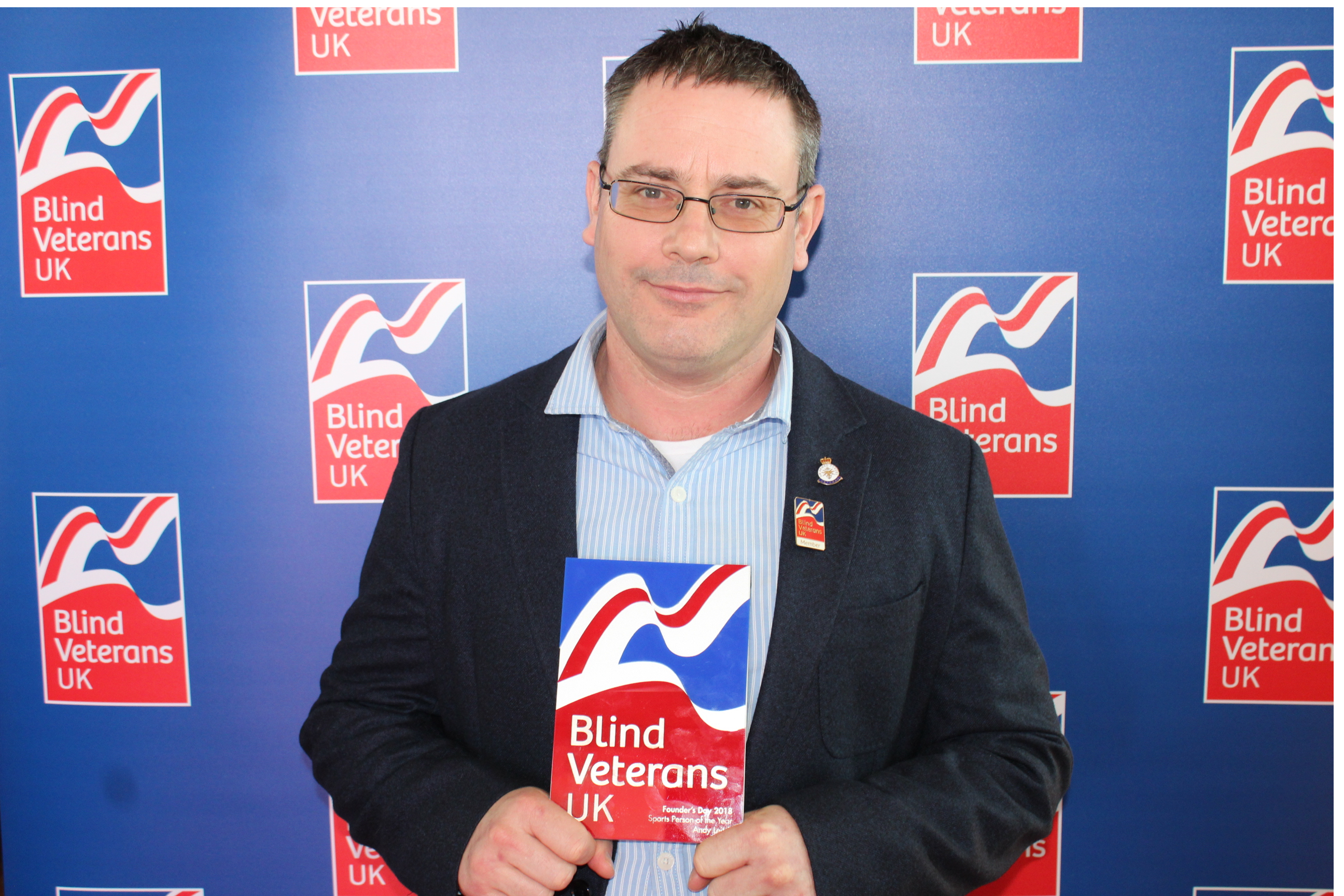 Blind veteran Andy is wearing a suit and holding his Founder's Day award. He is looking into camera and there is a Blind Veterans UK backdrop.