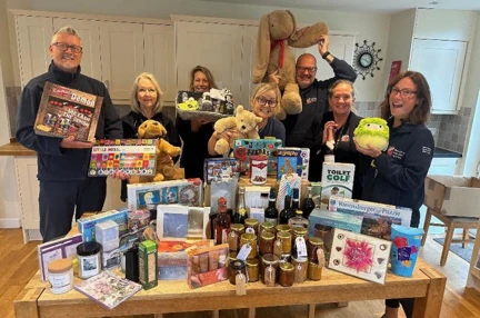 A group of Blind Veterans UK staff behind a table full of fundraising prizes in preparation for the first birthday fete and Open day on 7th September 2024
