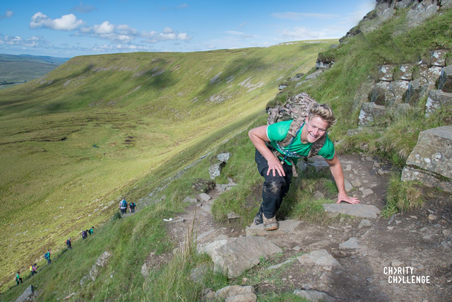 An event participant clambering up a steep narrow path on one of the peaks, below them other participants are following their footsteps.  The dramatic landscape of the Yorkshire Dales stretches off in the background below a blue sky dotted with clouds.