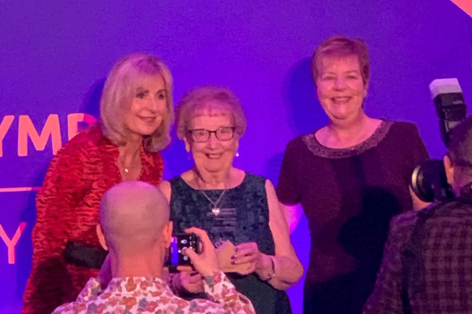 Gill is standing with the presenter and posing for photos by the photographers