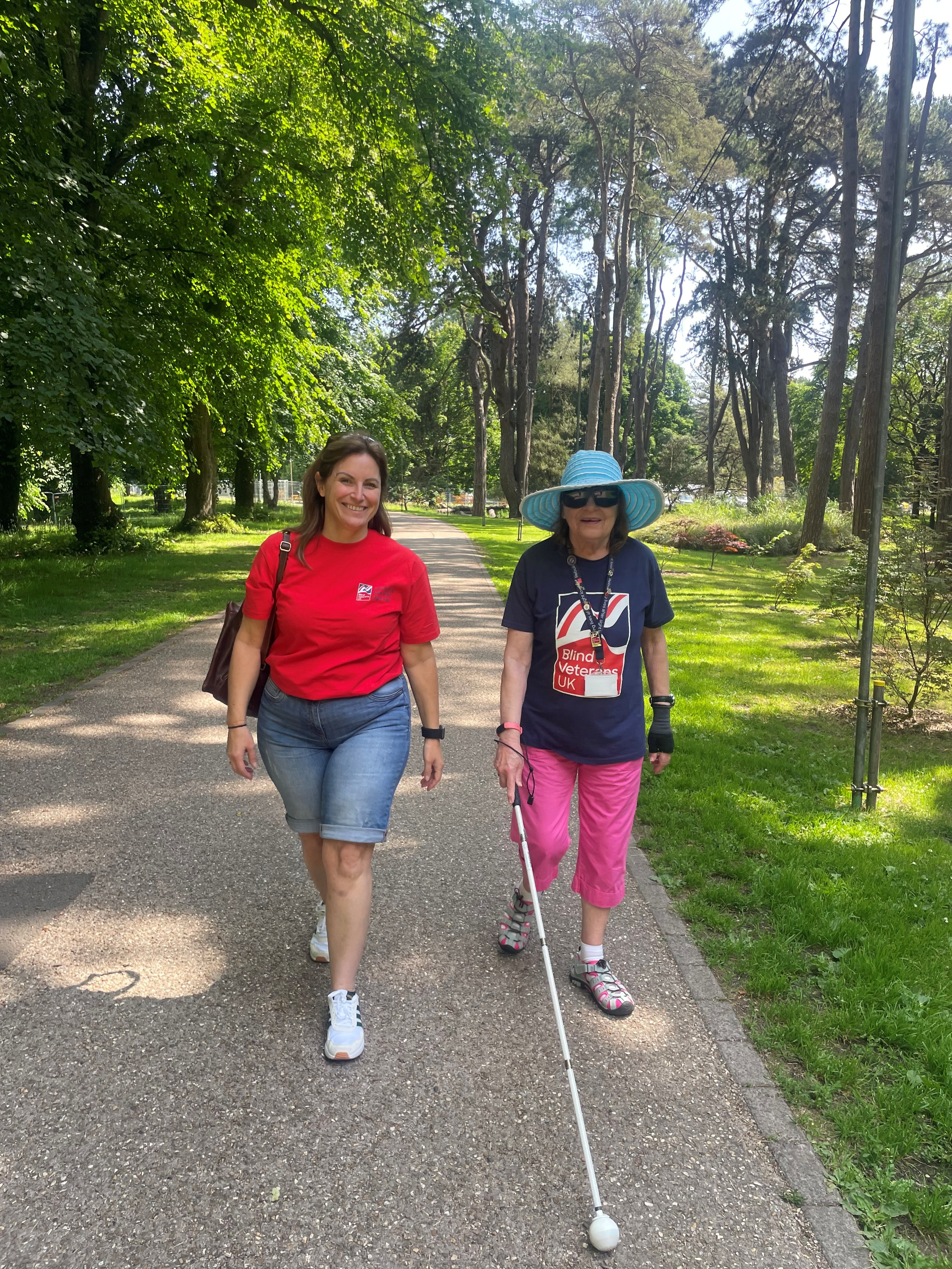 Beth and Noeline walk side by side along a tree lined pathway. Noeline is wearing a Blind Veterans UK t-shirt and using her cane.