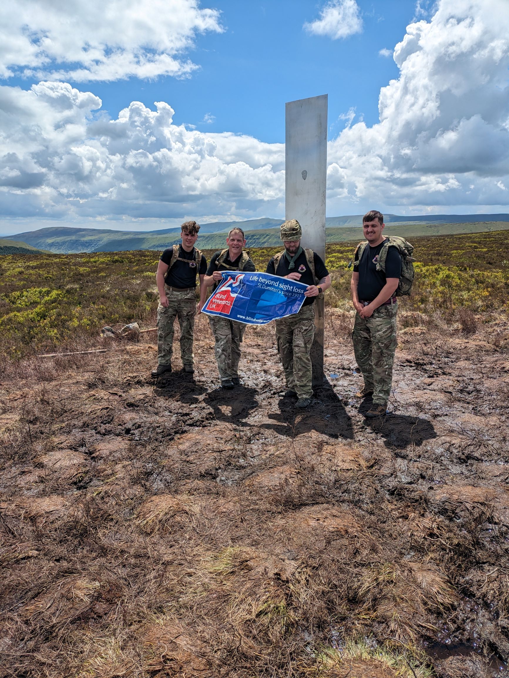 A group of four members of the Wrexham Army Careers Office all wearing camouflage trousers and carrying camouflage backpacks. They are stood with mountains behind them