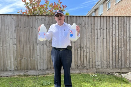 John stood in his garden wearing a Blind Veterans UK baseball cap and holding an empty four pint plastic milk carton in each hand with 'Every Penny Counts' labels attached