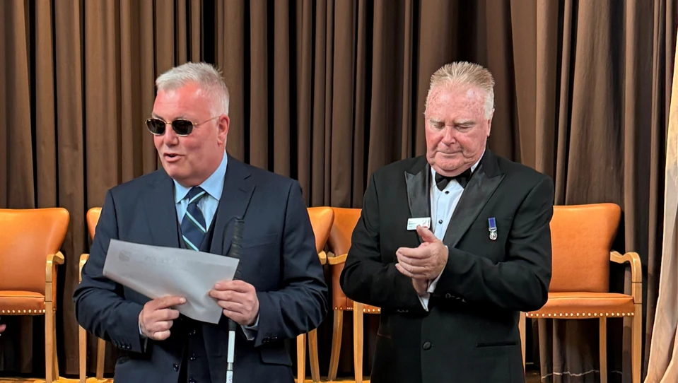 Blind veteran Niall is wearing dark sunglasses and holding a white cane as he receives his award. President Colin Williamson stands next to him clapping.