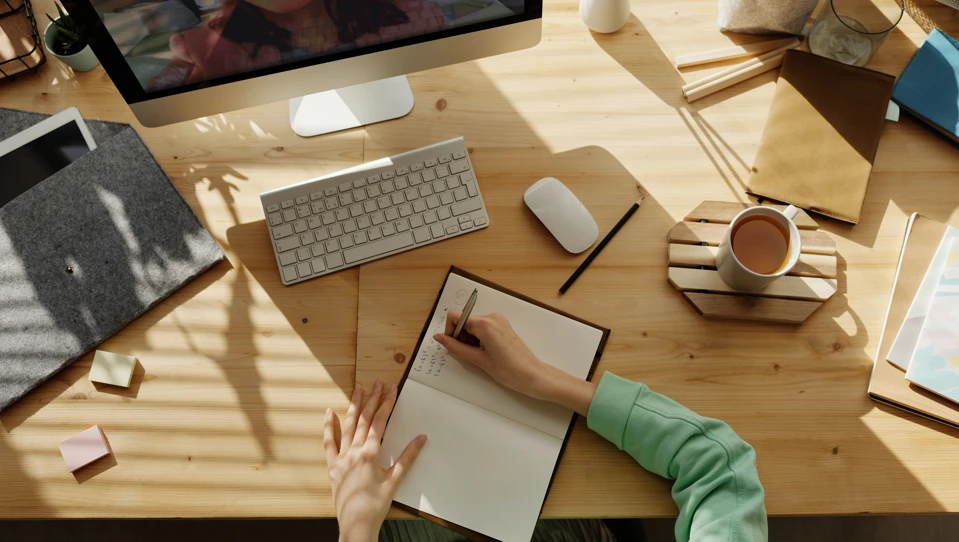 A woman sat at a desk writing in a notebook while on a computer video call
