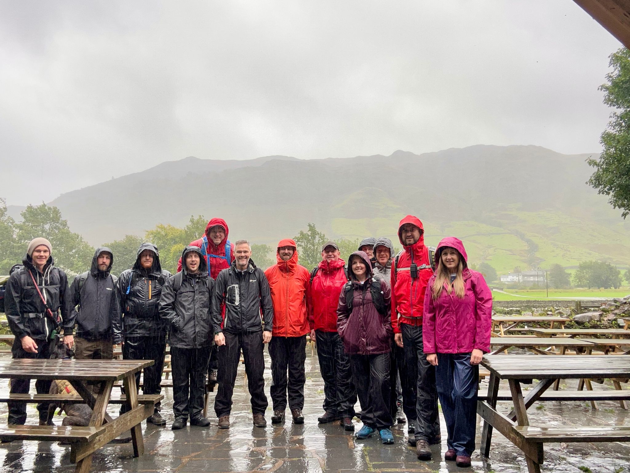 Group photo of the BOGE UK walkers stood around picnic benches with a mountain behind them. The group are all in rain coats with hoods up and are very wet