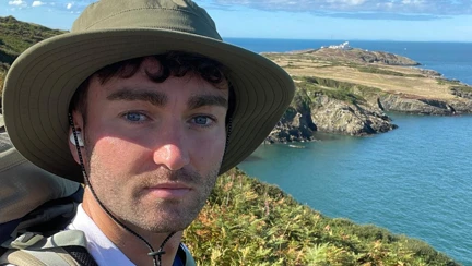 James wearing a Blind Veterans UK t shirt stood on the coastal path on a very sunny day with the sea in the background