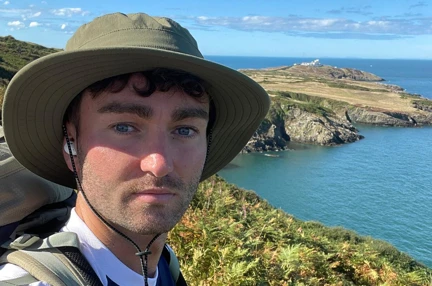 James wearing a Blind Veterans UK t shirt stood on the coastal path on a very sunny day with the sea in the background